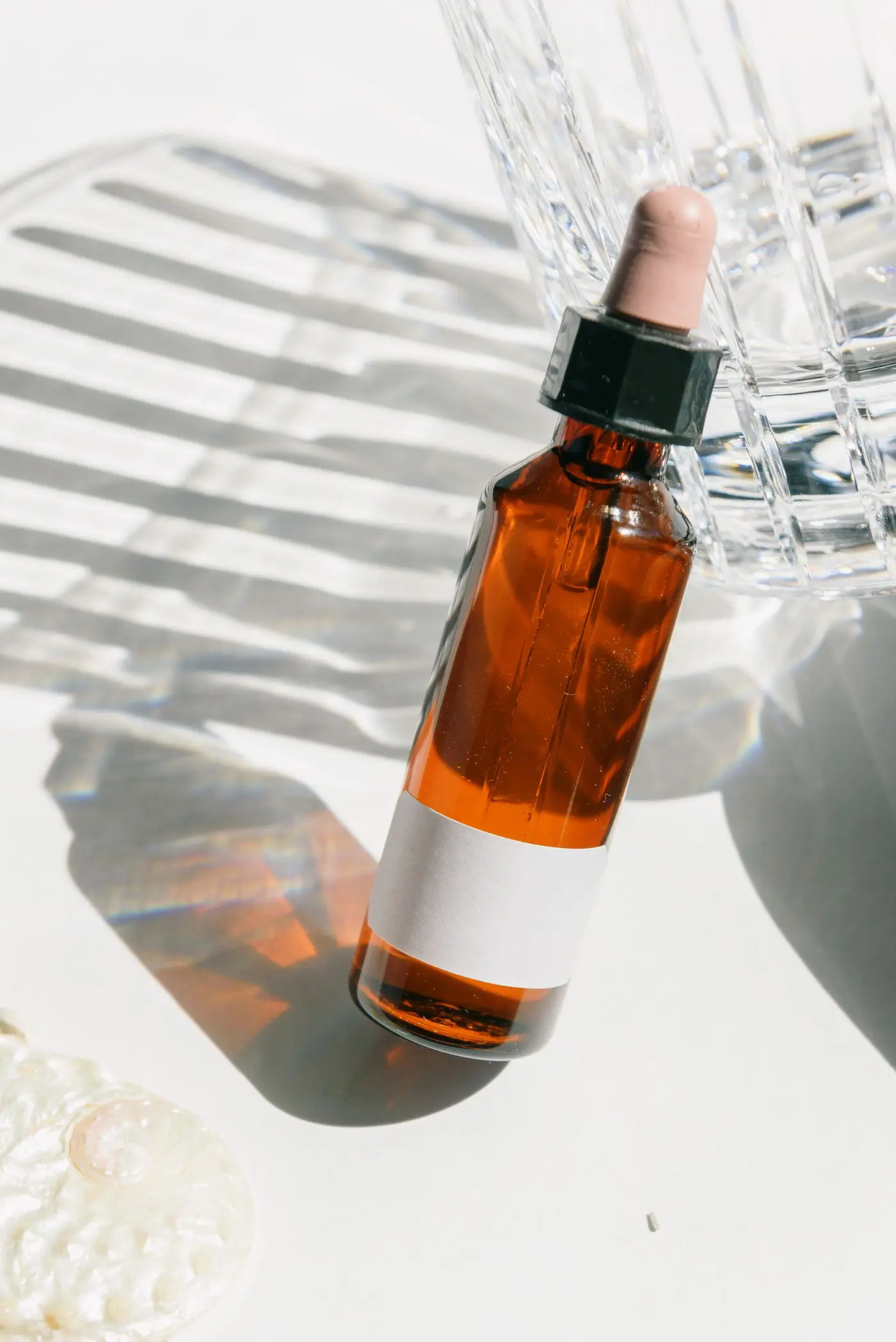 An amber dropper bottle beside a glass of water with striped shadows